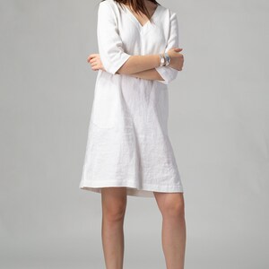 May include: A white linen dress with a v-neckline and 3/4 sleeves. The dress has a relaxed fit and a slightly A-line silhouette. The model is wearing the dress with brown espadrille sandals.