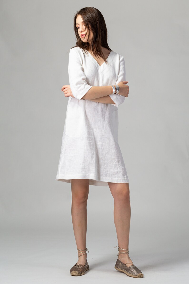 May include: A white linen dress with a v-neckline and 3/4 sleeves. The dress has a relaxed fit and a slightly A-line silhouette. The model is wearing the dress with brown espadrille sandals.
