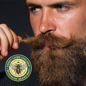 May include: A man with a beard and mustache is holding a strand of his beard. He is looking at the camera. The image includes a logo for Sweet Comb Chicago Mustache-Beard Wax. The logo is green and gold and features a bee with the text "Sweet Comb Chicago Rogers Park 60626 Made with Pure Natural Beeswax Mustache-Beard Wax."