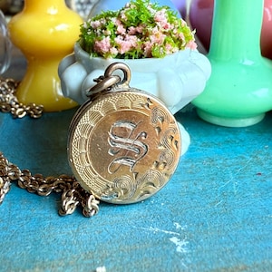 May include: A gold-colored locket necklace with an engraved design and the letter "S" in the center. The locket is round and attached to a delicate gold chain. The background includes colorful vases and a small white planter with pink and green foliage.