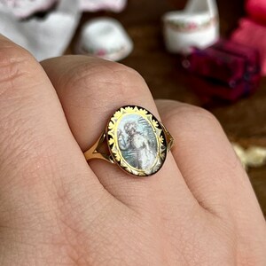 May include: A gold ring with an oval-shaped cameo featuring a painted portrait of a woman in a white dress. The cameo is surrounded by a black and gold filigree design.