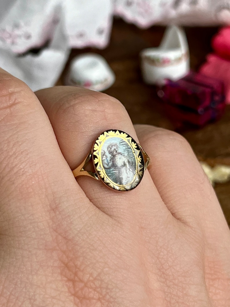 May include: A gold ring with an oval-shaped cameo featuring a painted portrait of a woman in a white dress. The cameo is surrounded by a black and gold filigree design.