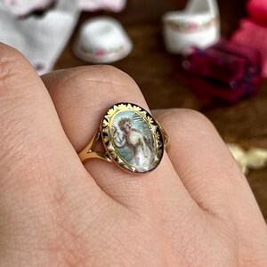 May include: A gold ring with a black and white oval cameo featuring a woman in a blue dress. The cameo is surrounded by a gold border with a floral design.