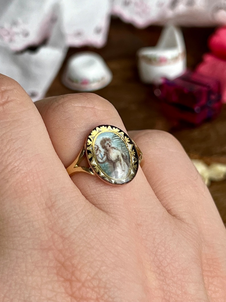 May include: A gold ring with a black and white oval cameo featuring a woman in a blue dress. The cameo is surrounded by a gold border with a floral design.