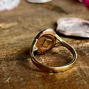 May include: A gold ring with a round, oval-shaped face. The face of the ring has a small, clear stone set in the centre. The ring is on a wooden surface.