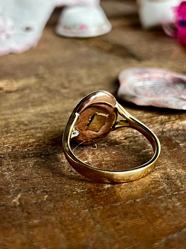 May include: A gold ring with a round, oval-shaped face. The face of the ring has a small, clear stone set in the centre. The ring is on a wooden surface.