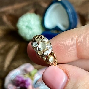 Puede incluir: Un anillo de oro con una piedra preciosa grande, clara y facetada rodeada de piedras preciosas más pequeñas, claras y facetadas. El anillo está engastado en una banda de oro.