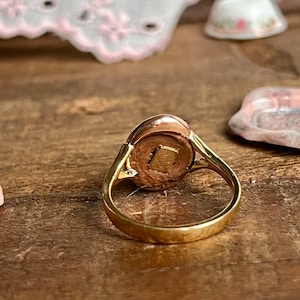 May include: A gold ring with a round, oval-shaped stone set in the centre. The stone is a light brown colour and has a smooth, polished surface.