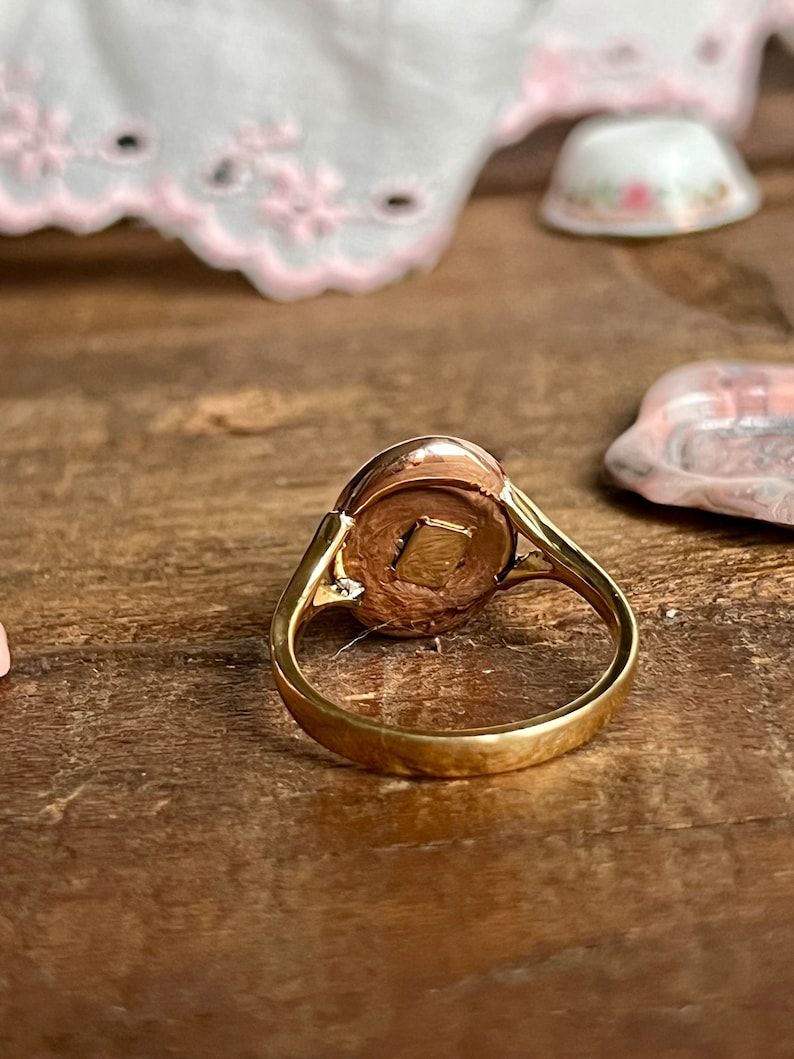 May include: A gold ring with a round, oval-shaped stone set in the centre. The stone is a light brown colour and has a smooth, polished surface.