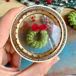 May include: A round, clear-domed ornament with a green crocheted wreath and a red bow. The ornament is trimmed with rhinestones and contains small decorative elements. The background includes fabric with red and green patterns.