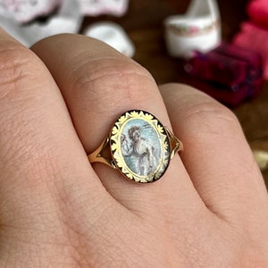May include: A gold ring with an oval-shaped cameo featuring a painted image of a woman in a white dress. The cameo is surrounded by a gold border with a floral design.