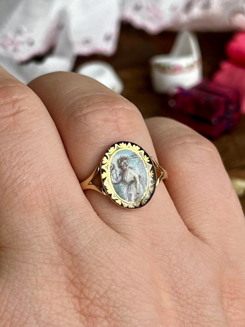 May include: A gold ring with an oval-shaped cameo featuring a painted image of a woman in a white dress. The cameo is surrounded by a gold border with a floral design.