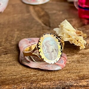 May include: A gold ring with an oval-shaped cameo featuring a detailed image of a person in a white dress. The cameo is surrounded by a black and gold border.