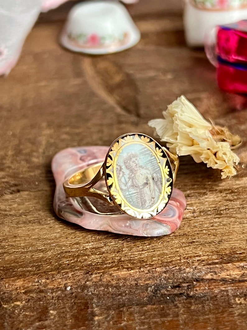 May include: A gold ring with an oval-shaped cameo featuring a detailed image of a person in a white dress. The cameo is surrounded by a black and gold border.