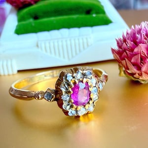 Op de afbeelding: Een gouden ring met een centrale roze edelsteen, omringd door kleinere witte en grijze stenen. De ring heeft een gedetailleerd, sierlijk ontwerp en ligt op een gouden oppervlak. Roze bloemen zijn op de achtergrond te zien.
