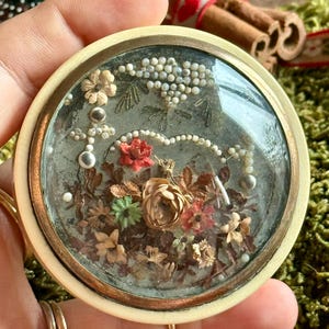 May include: A round, cream-coloured trinket box with a glass top. The box is filled with dried flowers and beads, creating a delicate and intricate design.