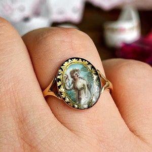 May include: A gold ring with a black and white oval cameo featuring a woman in a white dress. The cameo is surrounded by a gold and black border.