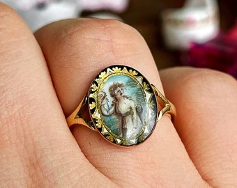 Puede incluir: Un anillo de oro con un camafeo ovalado en blanco y negro que representa a una mujer con un vestido blanco. El camafeo está rodeado por un borde dorado y negro.