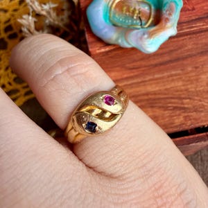 May include: A gold ring featuring a bypass design with two small square-cut gemstones, one vibrant pink and one deep blue, set into the polished metal band. The ring has a smooth, rounded surface that curves around the finger, showcasing the stones.