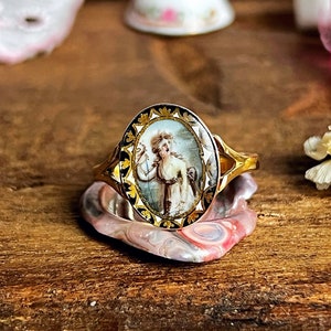 May include: A gold ring with an oval-shaped cameo featuring a painted portrait of a woman in a white dress. The cameo is surrounded by a black and gold border.