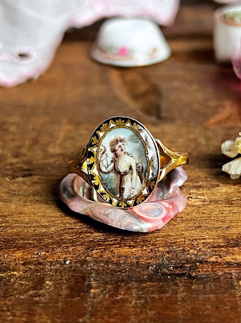 May include: A gold ring with an oval-shaped cameo featuring a painted portrait of a woman in a white dress. The cameo is surrounded by a black and gold border.