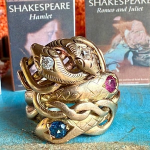 May include: A stack of gold-colored snake rings with gemstone eyes, including diamonds, a ruby, and a blue stone. The rings are intricately detailed and sit in front of miniature Shakespeare books.