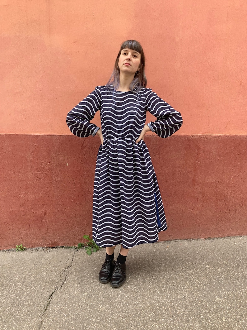 May include: A navy blue dress with a white wavy pattern. The dress has long sleeves and a flared skirt. The person in the image is wearing black shoes and socks. The background is a two-tone wall with a pinkish-red colour.