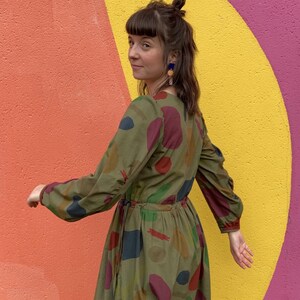 May include: An olive green dress with long sleeves and a colourful abstract pattern of red, blue, and yellow shapes. The dress has a cinched waist and is worn by a person with dark hair and blue earrings.