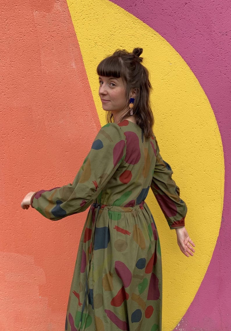 May include: An olive green dress with long sleeves and a colourful abstract pattern of red, blue, and yellow shapes. The dress has a cinched waist and is worn by a person with dark hair and blue earrings.