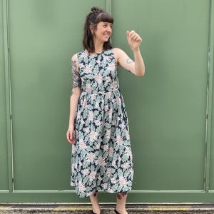 May include: A sleeveless dress with a floral pattern in navy, pale green, and cream. The dress has a fitted bodice and a flared skirt. The model is wearing black sandals. The background is a solid green wall.