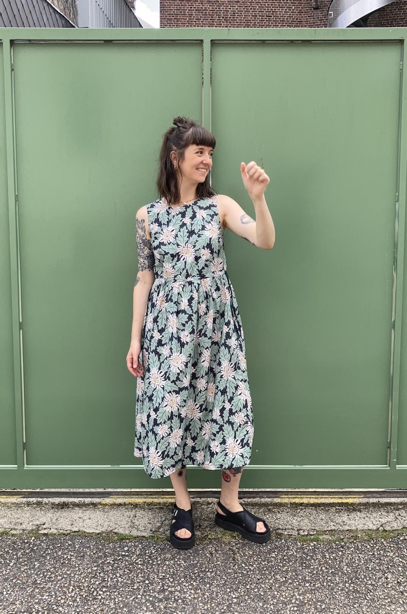 May include: A sleeveless dress with a floral pattern in navy, pale green, and cream. The dress has a fitted bodice and a flared skirt. The model is wearing black sandals. The background is a solid green wall.