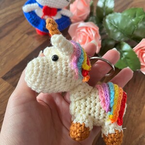 May include: A crocheted unicorn keychain with a rainbow mane and tail. The unicorn is white with brown hooves and a brown horn. The keychain is attached to a silver ring.