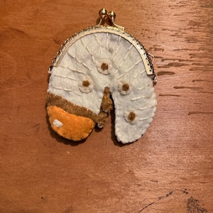May include: A semi-circular felt coin purse featuring a gold-toned metal kiss-lock clasp. The white felt body is decorated with embroidered white lines and small brown floral accents, while one side displays a contrasting orange and brown felt patch.