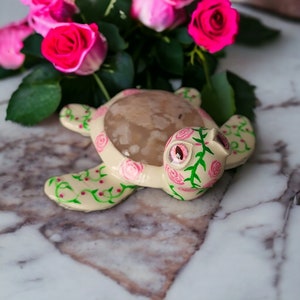 May include: A decorative turtle figurine with a cream-colored body and a brown shell. The turtle is adorned with pink roses and green vines. The background features pink roses and green leaves.