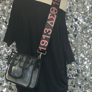 May include: A black clear tote bag with a black shoulder strap. The strap has red lettering that says "1913 ΔΣΘ 1913".