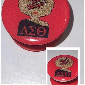 May include: Red phone grip with a black and gold design. The design features a woman with an afro and the text "Black Girl Magic". The letters "ΔΣΘ" are also featured on the grip.