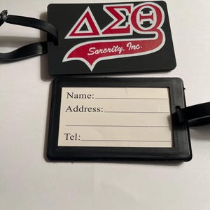 May include: Black luggage tag with a red and white Delta Sigma Theta Sorority, Inc. logo. The tag has a white label with spaces for a name, address, and telephone number.