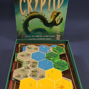 Puede incluir: Una caja verde con un tablero de juego en su interior. La caja tiene la palabra "CRYPTID" impresa en la parte delantera en letras blancas grandes. El tablero de juego está formado por fichas hexagonales con diferentes colores y diseños. Las fichas están dispuestas en un patrón circular.