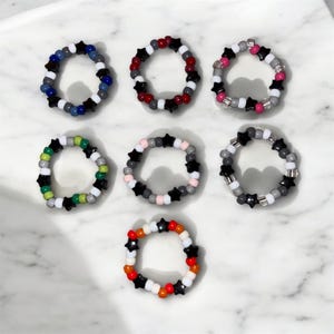 May include: Seven beaded rings in various color combinations. Each ring features black star-shaped beads interspersed with round beads in colors like white, gray, red, pink, and orange. The rings are arranged on a white surface.