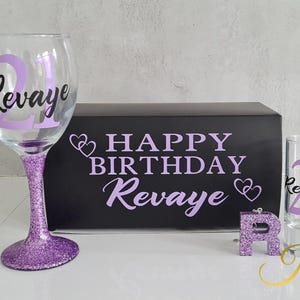 Może przedstawiać: Przezroczysty kieliszek do wina z fioletową brokatową nóżką oraz imieniem "Revaye" i liczbą "21" w kolorze fioletowym. Kieliszek do shotów z tym samym wzorem, czarne pudełko z napisem "Happy Birthday Revaye" w kolorze fioletowym i fioletowa brokatowa litera "R".