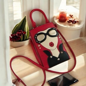 May include: A red handbag with a graphic of a woman's face, featuring large black-rimmed glasses, a red lip, and a black top. The bag has a matching red handle and shoulder strap, with silver studs. The bag is on a wooden surface.