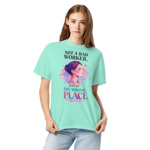 May include: A pale green t-shirt featuring the text "NOT A BAD WORKER. JUST IN THE WRONG PLACE." The design includes a woman's profile within a heart-shaped design with pink and purple watercolor effects.