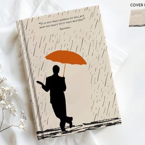 May include: A beige journal cover with a black silhouette of a person holding an orange umbrella. The background is a pattern of black rain drops. The quote "It is not what happens to you, but how you react to it that matters." by Epictetus is printed on the cover.