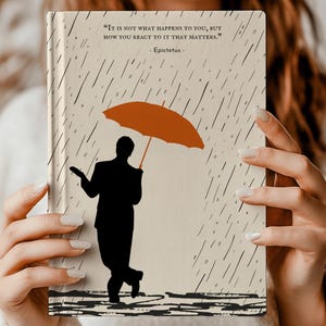 May include: A beige journal with a quote by Epictetus: "It is not what happens to you, but how you react to it that matters." The journal features a silhouette of a person holding an orange umbrella in a rain storm.