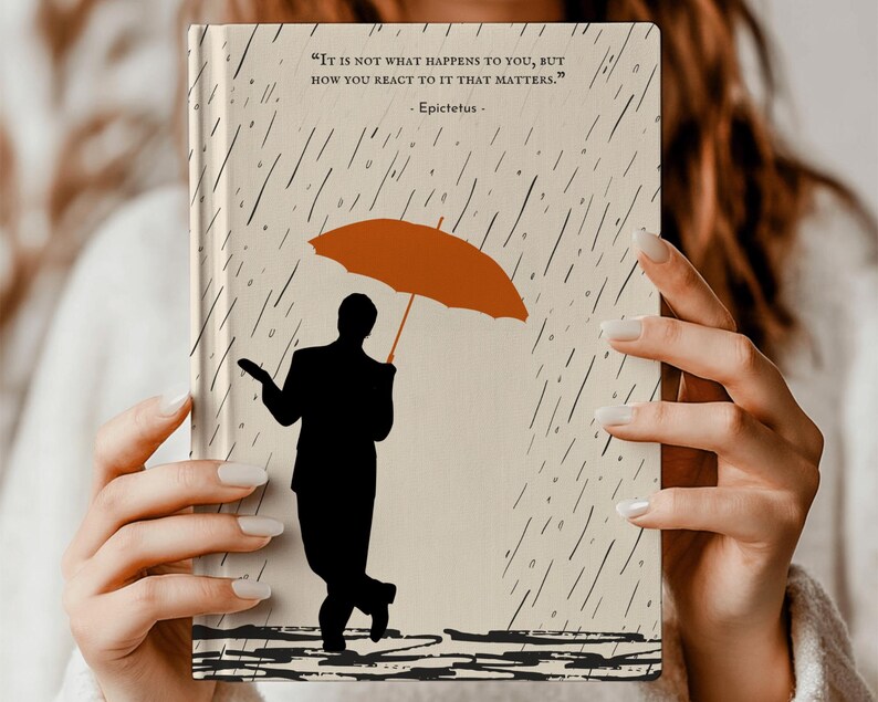 May include: A beige journal with a quote by Epictetus: "It is not what happens to you, but how you react to it that matters." The journal features a silhouette of a person holding an orange umbrella in a rain storm.