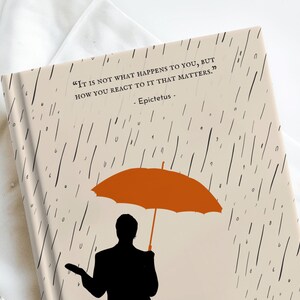 May include: A beige journal with a black and white rain pattern and a quote by Epictetus: "It is not what happens to you, but how you react to it that matters." A silhouette of a person holding an orange umbrella is in the lower right corner.