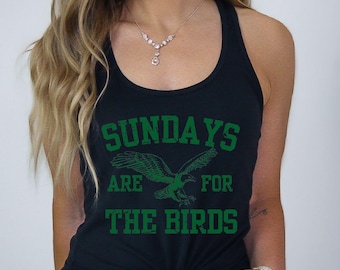 May include: A black racerback tank top featuring a green graphic of an eagle in flight centered between the text "SUNDAYS" above and "ARE FOR THE BIRDS" below. The design has a distressed, weathered texture and is printed in a bold, sans-serif font.