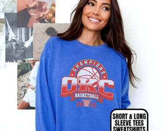 Może przedstawiać: Niebieska bluza z nadrukiem koszykówki z napisem "OKC Basketball Champions". Bluza z długim rękawem ma okrągły dekolt. Tekst "Flo Blue" znajduje się w lewym dolnym rogu, a "Short & Long Sleeve Tees Sweatshirts" po prawej stronie.