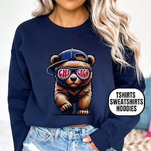 May include: Navy blue sweatshirt featuring a cartoon bear wearing a baseball cap and sunglasses with "CHI CAGO" text. The bear is walking forward. The sweatshirt has the text "TSHIRTS SWEATSHIRTS HOODIES" on the side.