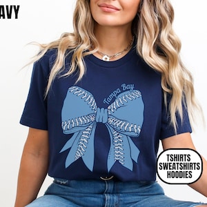 May include: Navy blue t-shirt featuring a light blue bow design with baseball stitching and the text "Tampa Bay". The shirt also has the text "Tshirts Sweatshirts Hoodies" in a white and black circle.